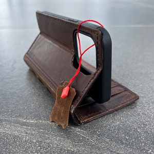 May include: Brown leather phone case with a black phone inside. The case is open, revealing a wallet-style design with card slots. A red string and a brown leather tag hang from the case.