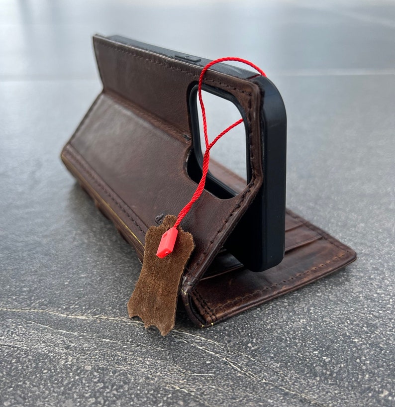 May include: Brown leather phone case with a black phone inside. The case is open, revealing a wallet-style design with card slots. A red string and a brown leather tag hang from the case.