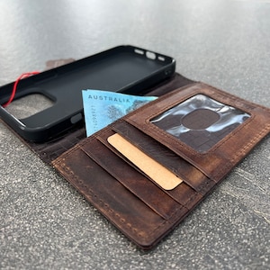 May include: A brown leather wallet with multiple card slots and a clear ID window. A black phone case and Australian currency are visible. The wallet is open, displaying its interior.