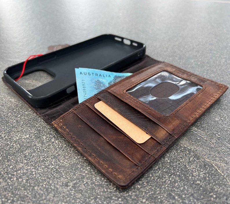 May include: A brown leather wallet with multiple card slots and a clear ID window. A black phone case and Australian currency are visible. The wallet is open, displaying its interior.