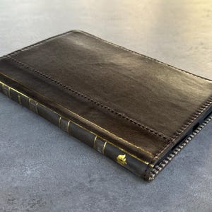 May include: A brown leather book-style case with gold accents. The case has a stitched design and a zipper closure. The spine features gold vertical lines and decorative elements. The case is designed to hold a device.