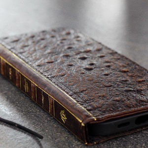 May include: A brown leather phone case designed to look like a vintage book. The case has a textured surface, gold accents, and a black interior. A black cord with a tassel is attached to the spine.