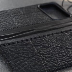 May include: A black leather phone case with a textured finish. The case has a wallet design with a flap and a strap. The case is designed to hold a phone and other items.