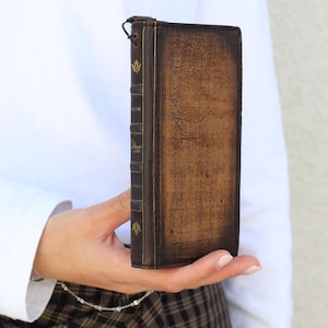 May include: A brown leather book-style wallet with gold accents and a decorative emblem. The wallet has a distressed appearance, with visible text on the spine. The wallet is held in a person's hand.
