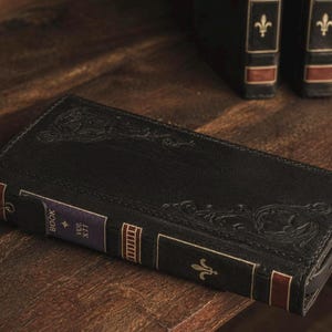 Op de afbeelding: Een zwarte lederen telefoonhoes ontworpen als een antiek boek. De kaft is voorzien van in reliëf aangebrachte florale krullen. De rug toont de tekst BOOK VOL XII met een gouden lelie-embleem en rode en paarse accenten op een donkere achtergrond.