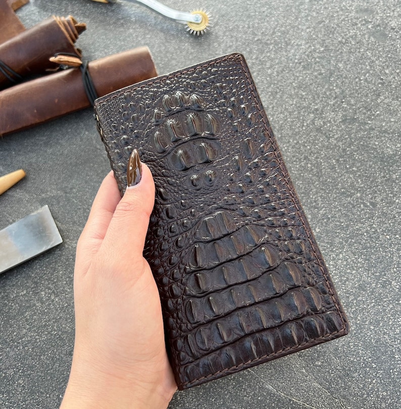 May include: A brown leather wallet with a crocodile embossed pattern. The wallet is closed and has a smooth, glossy finish.