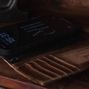 K&ouml;nnte beinhalten: Eine dunkelbraune Leder-Handyh&uuml;lle im Brieftaschenformat mit mehreren vertikalen Kartenf&auml;chern und einem transparenten Sichtfenster mit eingepr&auml;gtem Logo. Ein schwarzes Smartphone mit einer digitalen Uhrzeitanzeige von 18:31 liegt in der H&uuml;lle.