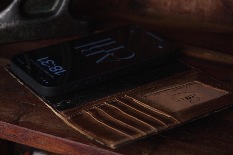K&ouml;nnte beinhalten: Eine dunkelbraune Leder-Handyh&uuml;lle im Brieftaschenformat mit mehreren vertikalen Kartenf&auml;chern und einem transparenten Sichtfenster mit eingepr&auml;gtem Logo. Ein schwarzes Smartphone mit einer digitalen Uhrzeitanzeige von 18:31 liegt in der H&uuml;lle.