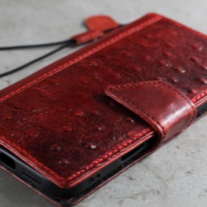May include: A red leather phone case with a textured finish and a magnetic clasp. The case features a matching red strap and is designed to protect a smartphone. The case is made of high-quality leather.
