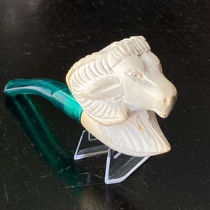 May include: A decorative bottle stopper featuring a ram's head carved from a light-colored material. The stopper has a green and teal handle. The ram's head is detailed with carved fur and facial features.