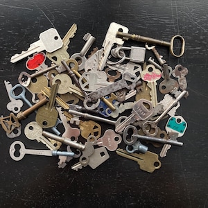 May include: A collection of various metal keys, in different sizes and shapes, scattered on a dark surface. Some keys have colored tags, and others have decorative designs. The keys are a mix of silver, brass, and other metals.