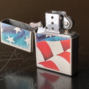 May include: A silver metal lighter with an open lid, displaying a design of the American flag. The lid features a blue background with white stars, while the body showcases red and white stripes. The lighter is on a dark surface.