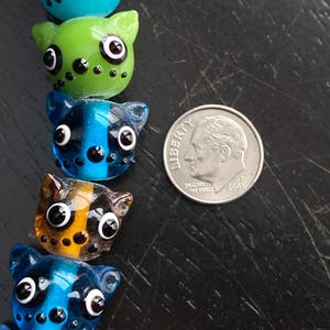 May include: A collection of colorful, cat-shaped glass beads with large eyes and small ears. The beads are in shades of blue, green, and orange, with black spots. A US dime is included for size reference.