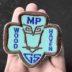 May include: A teal and brown embroidered patch with the text "MP WOOD HAVEN GS" and an owl design. The patch is in the shape of a trefoil, a symbol often associated with scouting organizations.