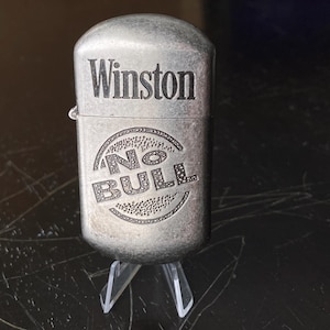 May include: A silver-toned Winston lighter with the words "Winston" and "NO BULL" engraved on the front. The lighter is displayed on a clear plastic stand, showcasing its design and brand.