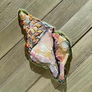 May include: A mosaic seashell sculpture with a variety of colors, including pink, yellow, orange, and white. The mosaic is made of small, irregularly shaped pieces. The sculpture is set against a light brown wooden background.