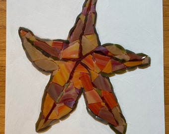 May include: A mosaic starfish art piece crafted from various shades of orange, red, yellow, and brown glass shards. The starfish is centered on a rectangular white board with a textured surface, featuring dark grout lines between the glass pieces.