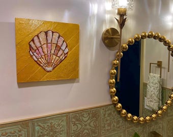 May include: A square wall art piece featuring a textured gold background with a central seashell illustration in shades of pink, white, and brown. The artwork is displayed on a wall next to a round gold beaded mirror and a gold wall sconce light.