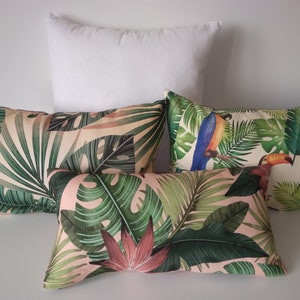 May include: A collection of decorative throw pillows with tropical leaf and toucan bird patterns. The pillows feature a variety of green leaves and pink flowers on a light pink background. One solid white pillow is also present.