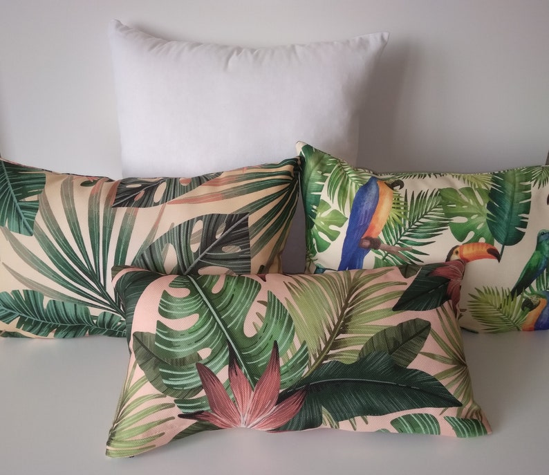 May include: A collection of decorative throw pillows with tropical leaf and toucan bird patterns. The pillows feature a variety of green leaves and pink flowers on a light pink background. One solid white pillow is also present.