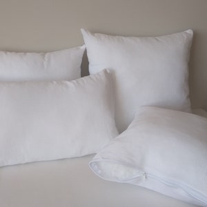 May include: A collection of four white pillows in various shapes and sizes. The pillows include a rectangular lumbar pillow, a square pillow, and two other square pillows. One pillow has a visible zipper.