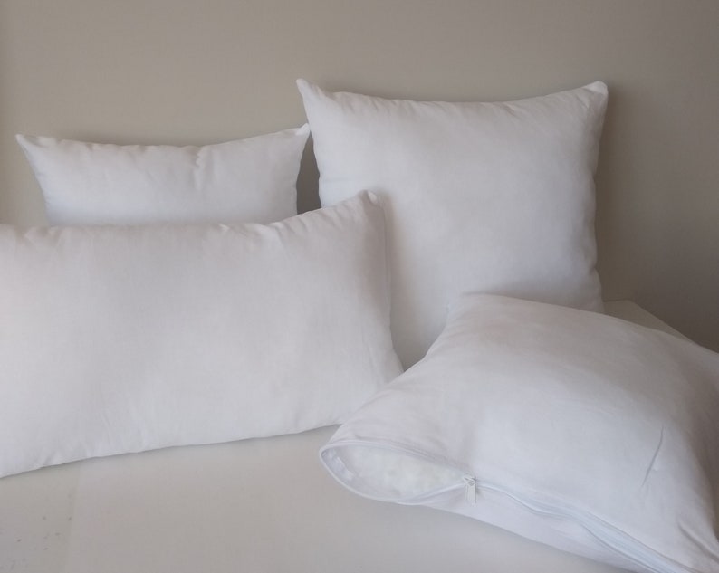 May include: A collection of four white pillows in various shapes and sizes. The pillows include a rectangular lumbar pillow, a square pillow, and two other square pillows. One pillow has a visible zipper.