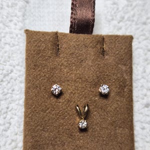 May include: A jewelry display featuring a brown suede-like card with a brown ribbon. The card holds a pair of stud earrings and a pendant, all with clear, round gemstones set in gold-colored metal.