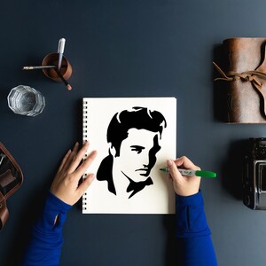 Puede incluir: Las manos de una persona dibujando una silueta en blanco y negro de Elvis Presley en una hoja de papel blanca en un cuaderno de espiral. La persona lleva una camisa azul de manga larga y est&aacute; usando un marcador verde.