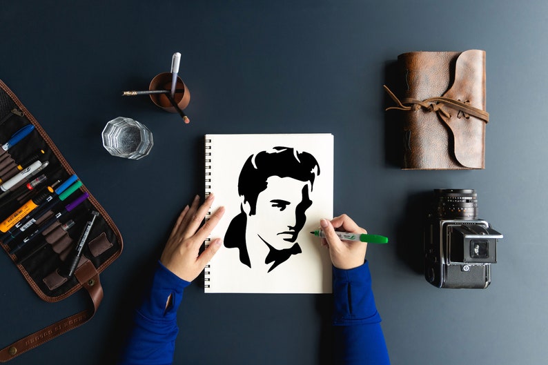 Puede incluir: Las manos de una persona dibujando una silueta en blanco y negro de Elvis Presley en una hoja de papel blanca en un cuaderno de espiral. La persona lleva una camisa azul de manga larga y est&aacute; usando un marcador verde.