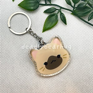 Può includere: Un portachiavi a forma di gatto beige con un'espressione triste. Il portachiavi è in acrilico trasparente e ha un anello in metallo argentato.