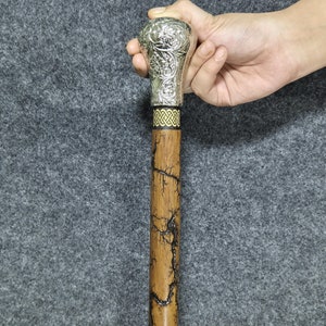 Può includere: Un bastone da passeggio in legno con un manico color argento che presenta un intricato disegno floreale. Il manico ha una fascia nera e dorata intorno. Il bastone ha una finitura marrone scuro con un effetto craquelé.