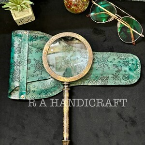 Puede incluir: Una lupa vintage con mango de lat&oacute;n y estuche de cuero. El estuche est&aacute; decorado con un tema n&aacute;utico y la lupa tiene la inscripci&oacute;n "Henry Hughes & Son Ltd. London 1921".