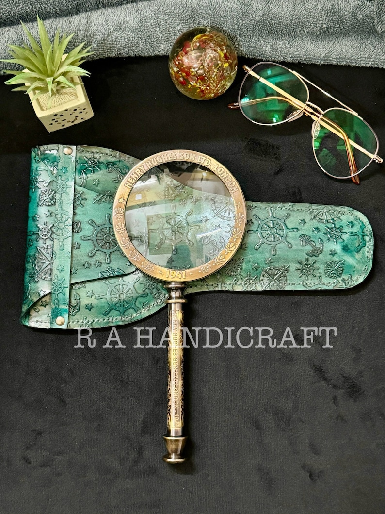 Puede incluir: Una lupa vintage con mango de lat&oacute;n y estuche de cuero. El estuche est&aacute; decorado con un tema n&aacute;utico y la lupa tiene la inscripci&oacute;n "Henry Hughes & Son Ltd. London 1921".