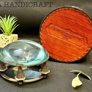 May include: A magnifying glass with a round, clear glass lens and a metal base with decorative feet. The magnifying glass is sitting on a brown surface. The magnifying glass is in a brown leather case with the text "HENRY HUGHES & SONS LTD LONDON 1949" embossed on the case.