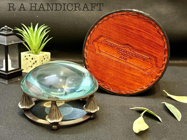 May include: A magnifying glass with a round, clear glass lens and a metal base with decorative feet. The magnifying glass is sitting on a brown surface. The magnifying glass is in a brown leather case with the text "HENRY HUGHES & SONS LTD LONDON 1949" embossed on the case.