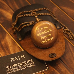 May include: A brass-colored compass pendant with the inscription "For our adventure forever and ever" on a leather case. The pendant hangs on a chain. A business card is also visible.