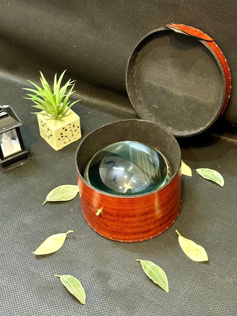 May include: A wooden box with a brass latch, containing a clear glass magnifying sphere. The box is lined with black felt and has a brown wood grain finish. The box is sitting on a black surface with green leaves scattered around it.