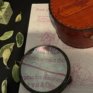 May include: A magnifying glass with a brass frame and a clear glass lens. The lens is magnifying a page of a book with the text "own to make the these first discover was & Chin" visible through the lens.