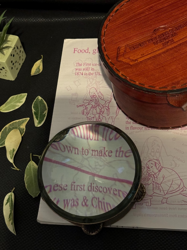 May include: A magnifying glass with a brass frame and a clear glass lens. The lens is magnifying a page of a book with the text "own to make the these first discover was & Chin" visible through the lens.