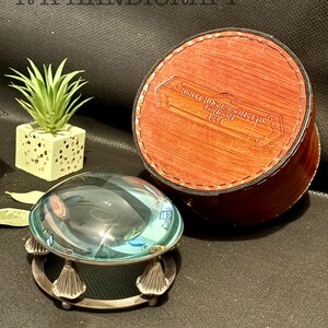 May include: A magnifying glass with a silver base and a clear glass lens. The magnifying glass is sitting on a dark surface. A brown leather box with the text "HENRY HILES SONS LTD LONDON 1921" is in the background.