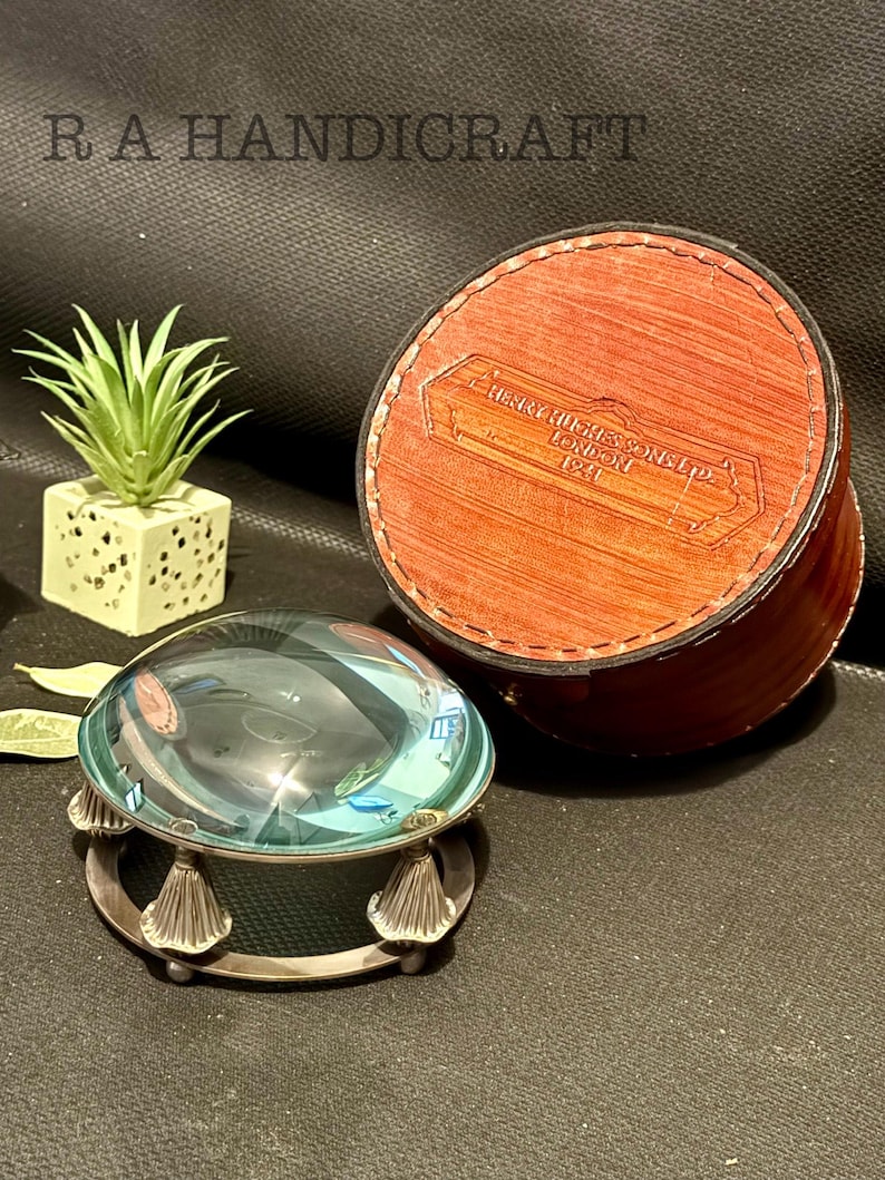 May include: A magnifying glass with a silver base and a clear glass lens. The magnifying glass is sitting on a dark surface. A brown leather box with the text "HENRY HILES SONS LTD LONDON 1921" is in the background.