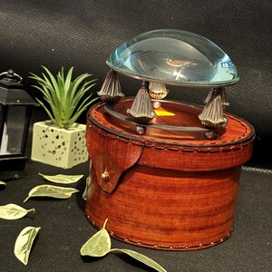 May include: A magnifying glass with a round, clear glass dome sits on top of a brown leather box with decorative stitching. The box has a gold-colored metal accent on the side.