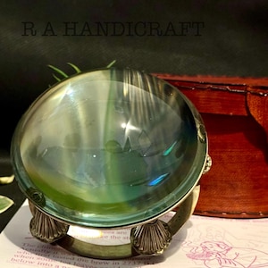 May include: A silver-colored magnifying glass with a round, clear glass lens. The lens is reflecting a green and blue pattern. The magnifying glass is sitting on a book with text.