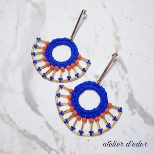 Puede incluir: Un par de pendientes hechos a mano con un diseño en forma de abanico. Los pendientes presentan un círculo azul tejido a ganchillo, cuentas de color coral y detalles dorados. Los pendientes tienen un poste dorado. El texto "atelier d'eder" está en la esquina inferior derecha.