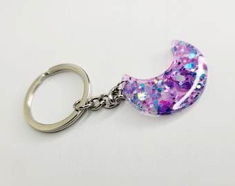 Può includere: Un portachiavi argentato con un ciondolo a forma di mezzaluna. Il ciondolo è pieno di glitter rosa, viola e blu.