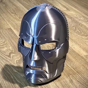 May include: A metallic silver full-face mask featuring a smooth, contoured surface with raised brow ridges, a prominent nose bridge, and horizontal eye openings. The design includes small circular rivet details along the edges and a segmented jawline.