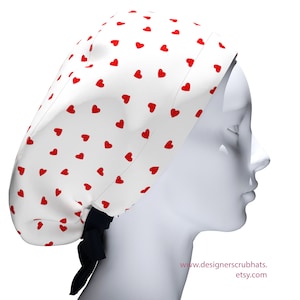 May include: A white scrub hat with a pattern of small red hearts. The hat is tied at the back with a black ribbon. The text "designerscrubhats.etsy.com" is visible on the lower right.