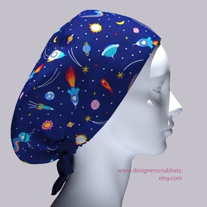 May include: A blue surgical scrub hat with a colorful space-themed print featuring rockets, planets, stars, and comets. The hat has a tie at the back.