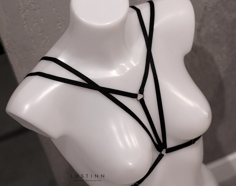 May include: Black lingerie harness with adjustable straps and metal rings. The harness is made of a soft, stretchy material and is designed to be worn over the chest.