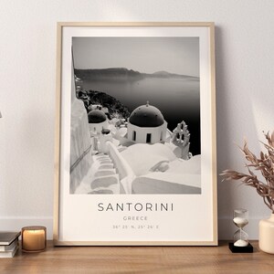 May include: Black and white photograph of Santorini, Greece. The image shows a white building with a dome roof and a staircase leading up to it. The sea and a small island are visible in the background. The text "SANTORINI, GREECE, 36&deg; 25' N, 25&deg; 26' E" is printed below the image.
